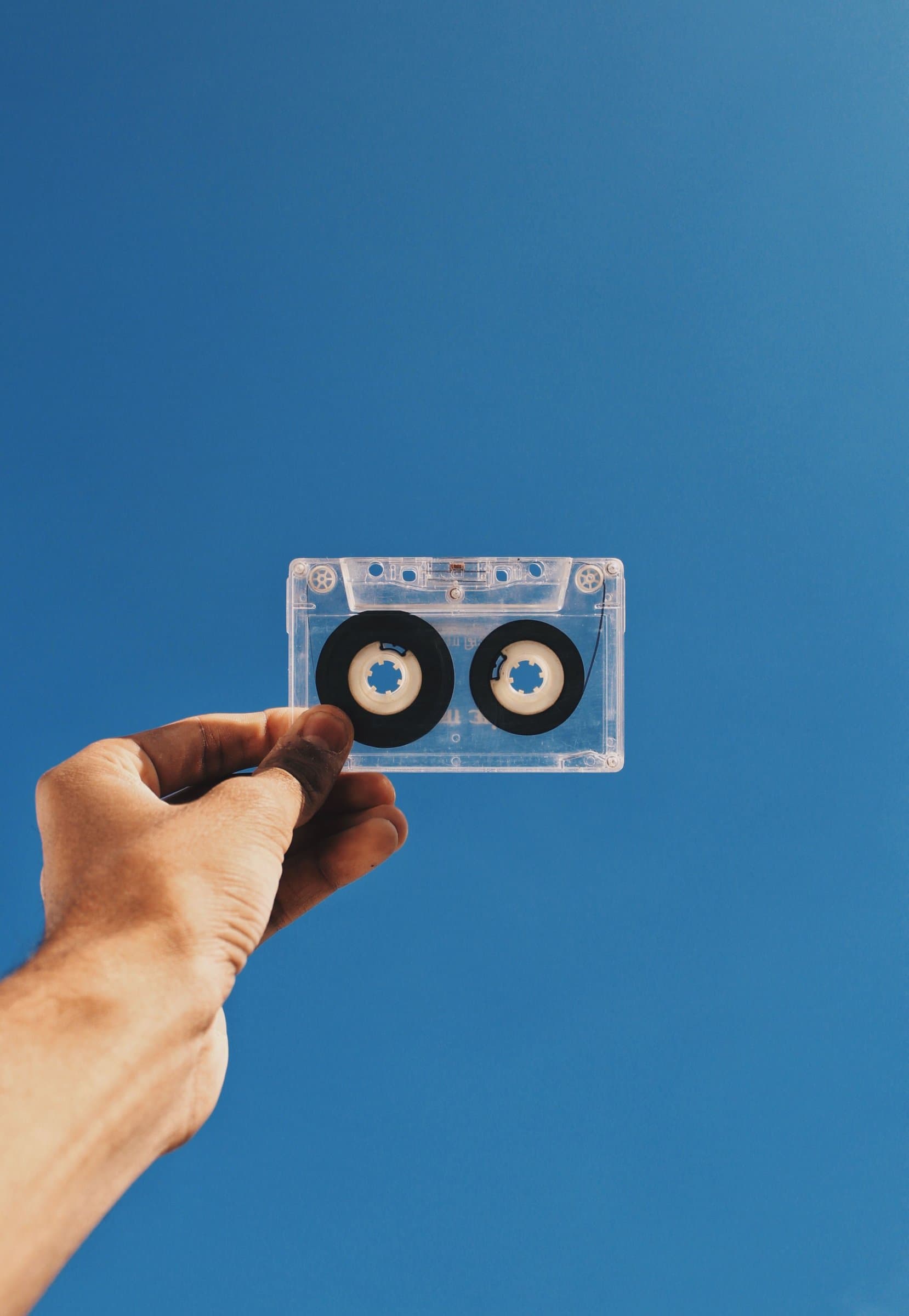 A cassette tape held up to a clear blue sky, its clear plastic shell catching the light.