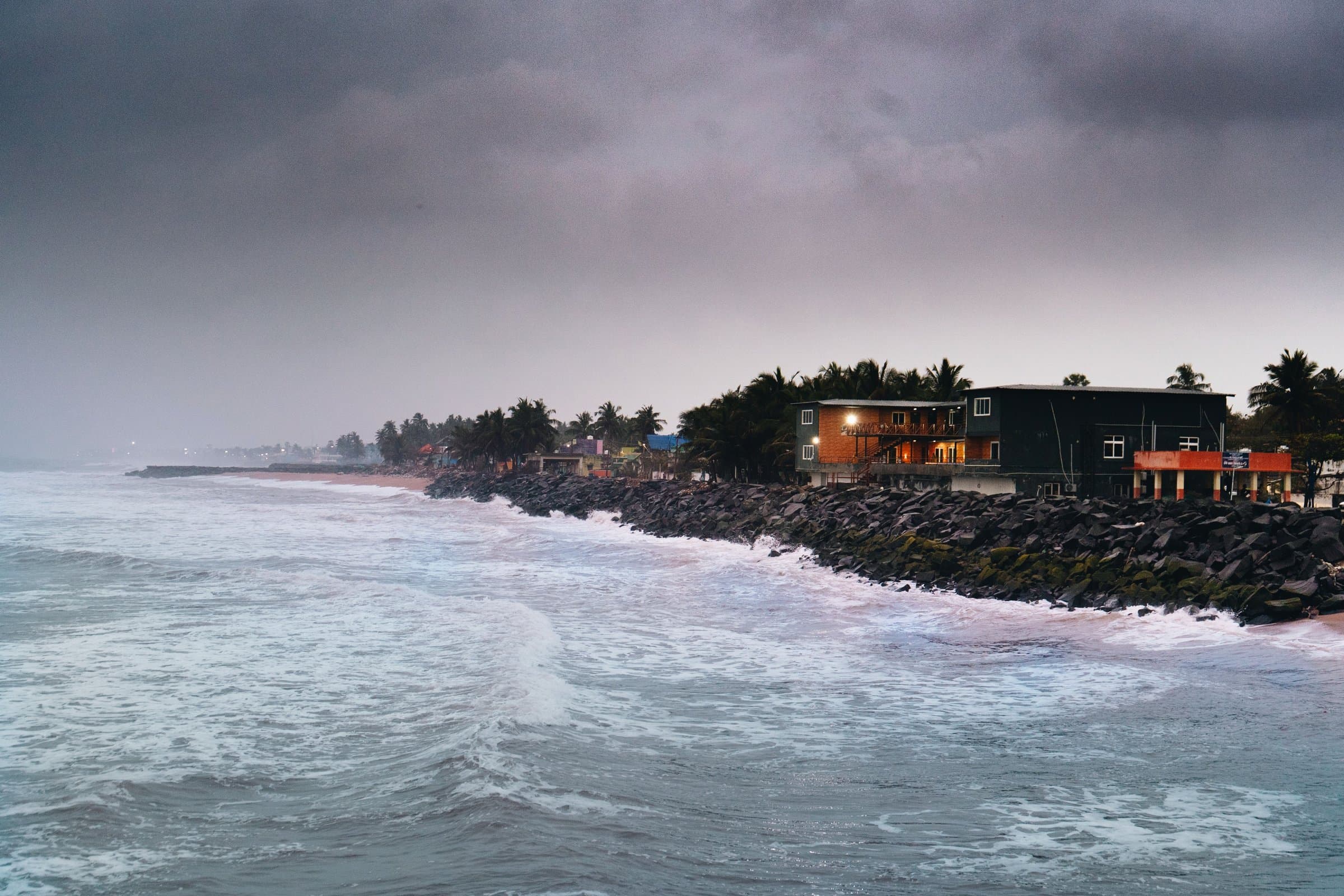 Storm clouds gather over a rocky coastline at dusk, waves breaking against black boulders.