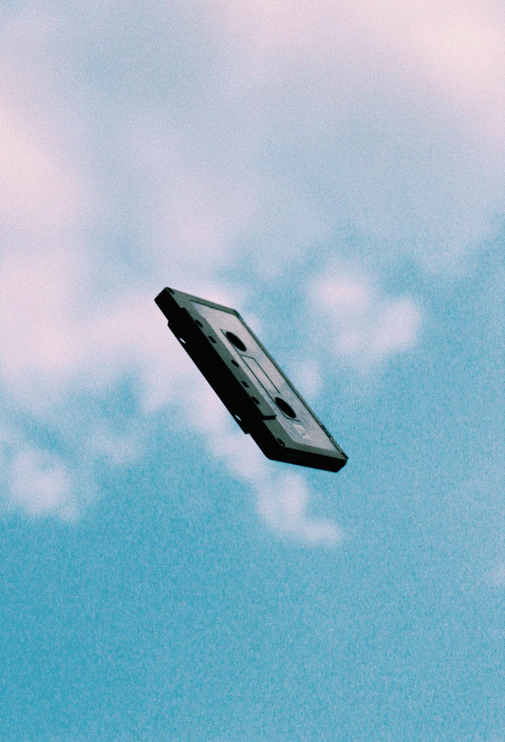 A cassette tape frozen mid-air against a grainy, pastel blue sky.
