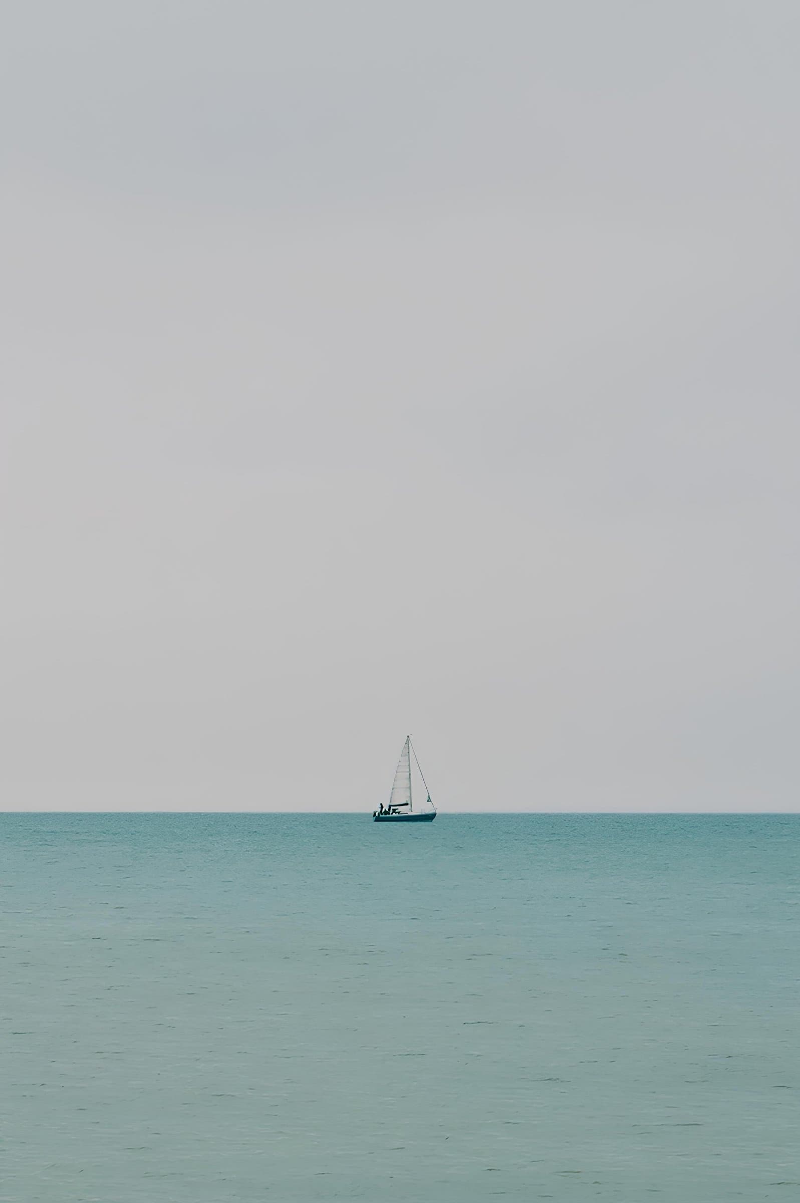 A small sailboat drifts alone on a flat, hazy sea beneath an overcast sky.