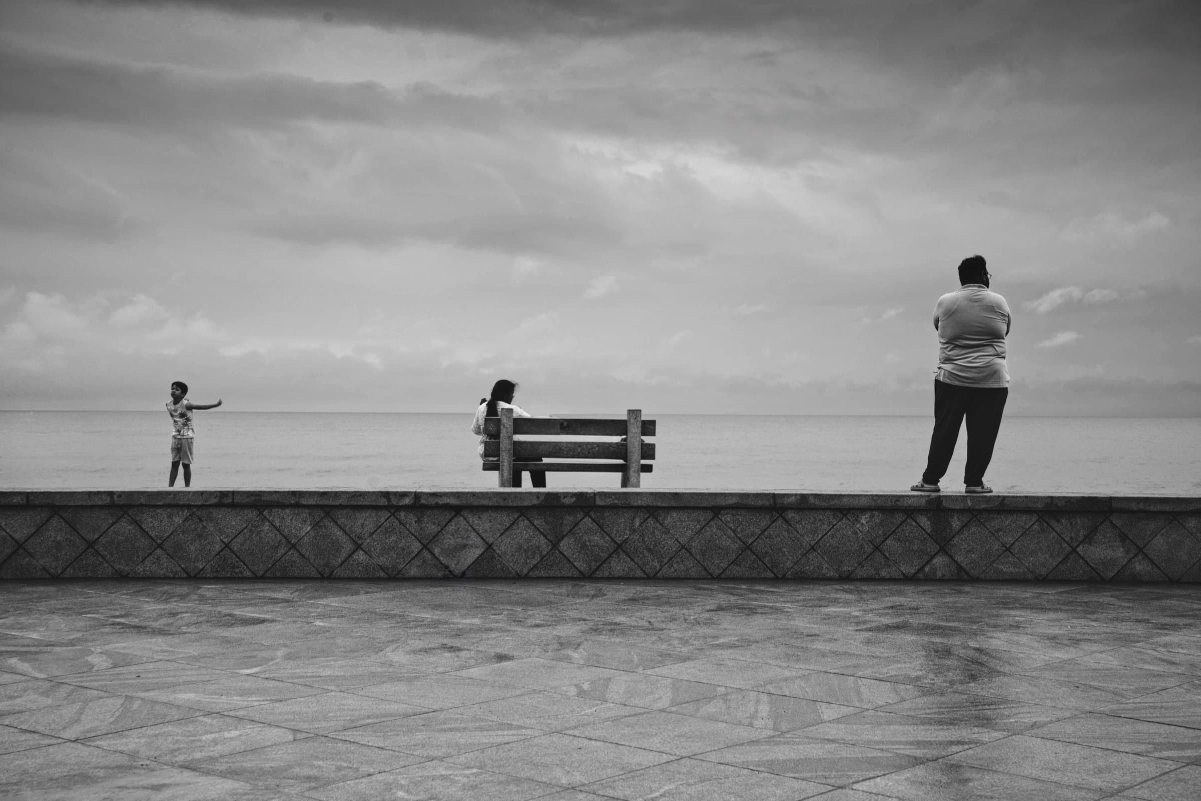 Three silhouetted figures along a seafront promenade wall, black and white, sea beyond.