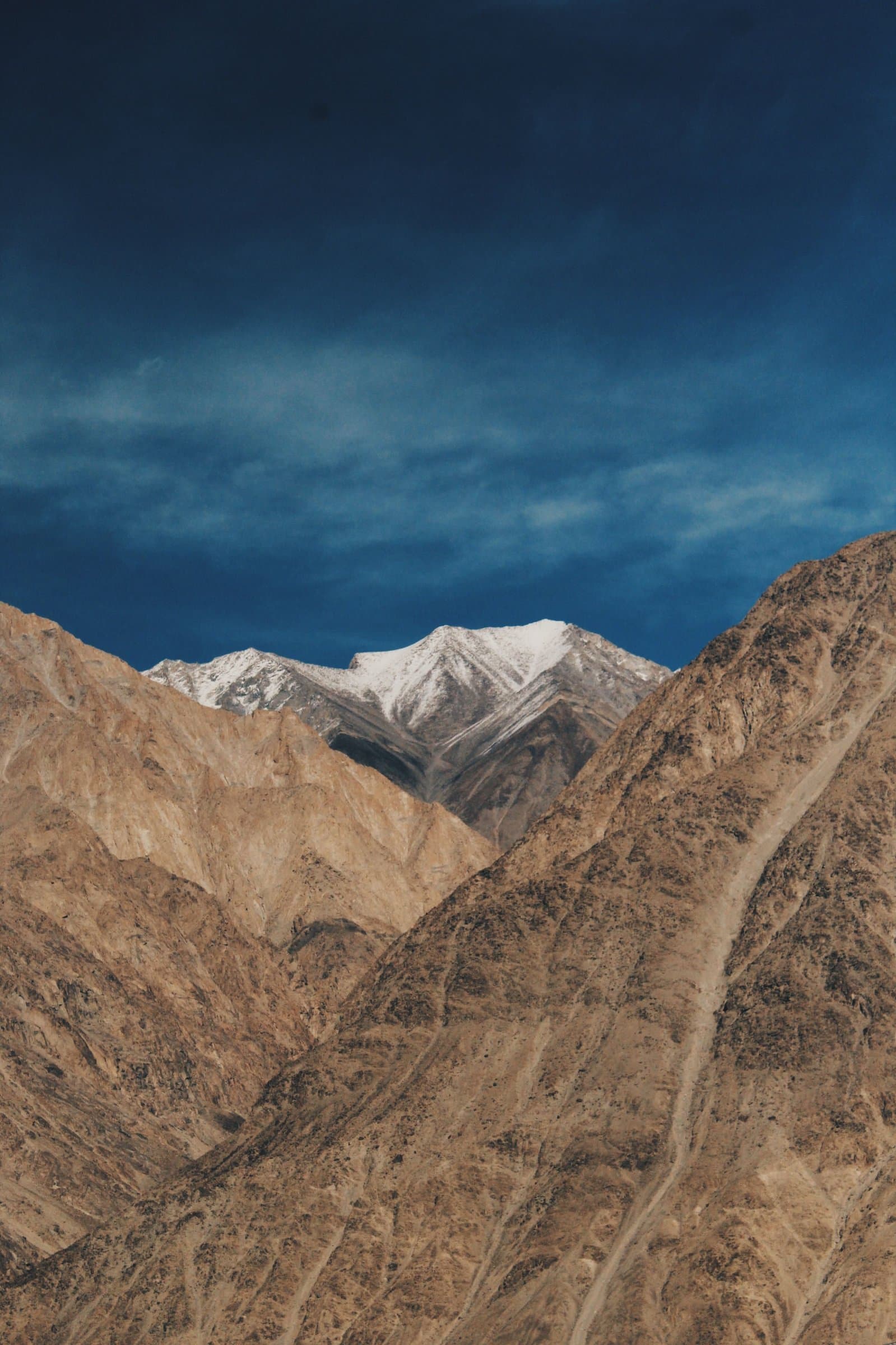 A snow-capped peak rising behind bare, folded mountains under a darkening sky.